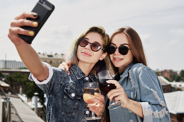 Two young smiling women drinking beer, taking selfie and having fun on the roof outdoors - Foto, afbeelding