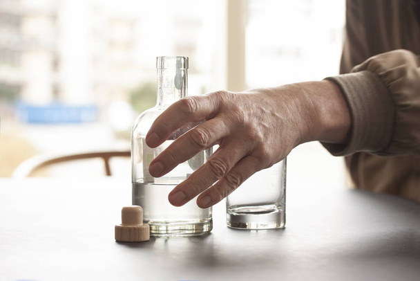 Man reaches out for a bottle of alcohol and an empty glass by the side at a table in home or bar environment. Selective focus on hand and bottle. Close up image - Photo, image