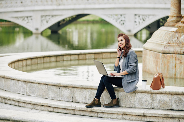 Внештатная симпатичная улыбающаяся женщина сидит у фонтана, работает на ноутбуке и звонит по телефону клиенту
 - Фото, изображение