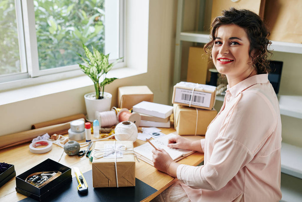 Ofisinde çalışan güzel, gülümseyen bir kadın paketleri paketliyor ve planlamacıyla yazıyor. - Fotoğraf, Görsel