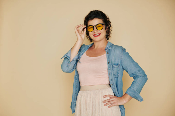 Portrait of pretty smiling woman putting on glasses with yellow lens and looking at camera - Photo, Image
