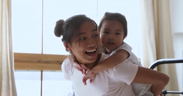 Smiling asian mum piggybacking child daughter playing laughing in bed - Video, Çekim