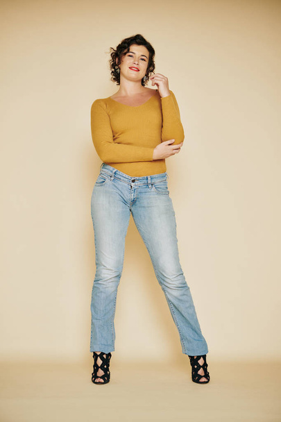 Красивая улыбающаяся молодая женщина в джинсах и горчичном топе с длинными рукавами позирует у бежевой стены
 - Фото, изображение