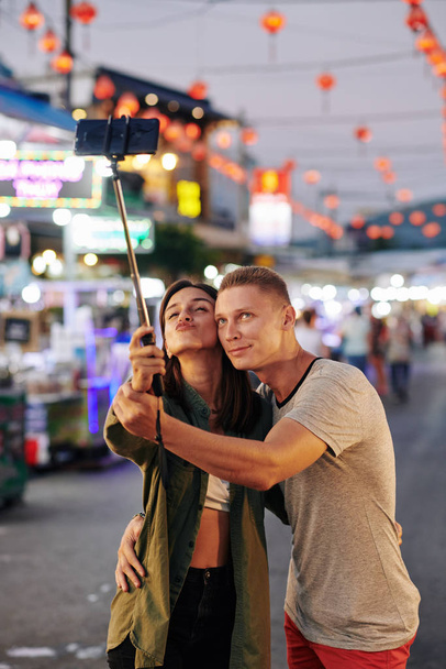 Счастливая молодая пара влюблённая делает селфи на смартфоне во время прогулки по азиатскому уличному рынку вечером
 - Фото, изображение