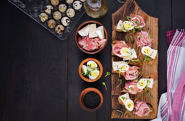 Bruschetta with prosciutto/jamon traditional Italian antipasto. Delicious snack with bread, brie cheese and quails eggs. Health food, tapas. Top view, copy space - Photo, Image