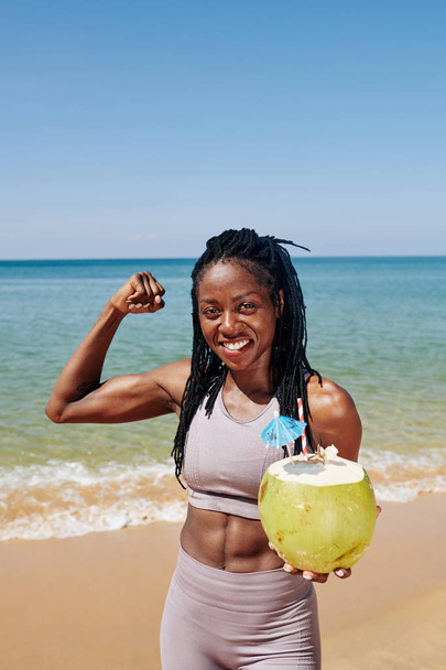 Positivo in forma giovane donna nera bere cocktail di cocco e mostrando muscoli bicipiti
 - Foto, immagini