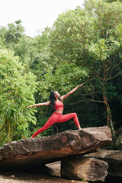 Joven deportista negra delgada disfrutando practicando yoga sobre roca grande en el bosque
 - Foto, Imagen
