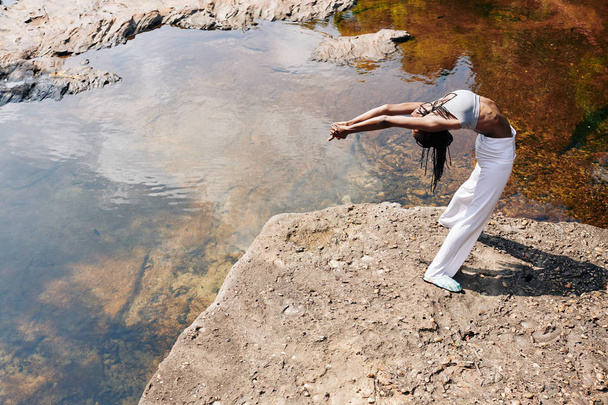 zwarte jonge vrouw in sportkleding doen terug buigen wanneer staan op rots door de rivier - Foto, afbeelding