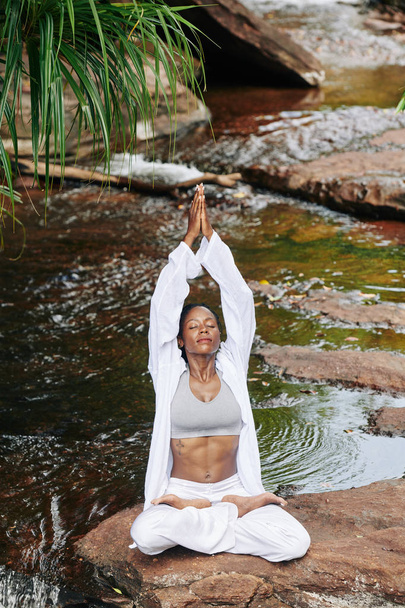 Zdolna młoda kobieta w bawełnianym garniturze podnosi ręce w namaste gesty medytując w pięknym miejscu nad rzeką - Zdjęcie, obraz