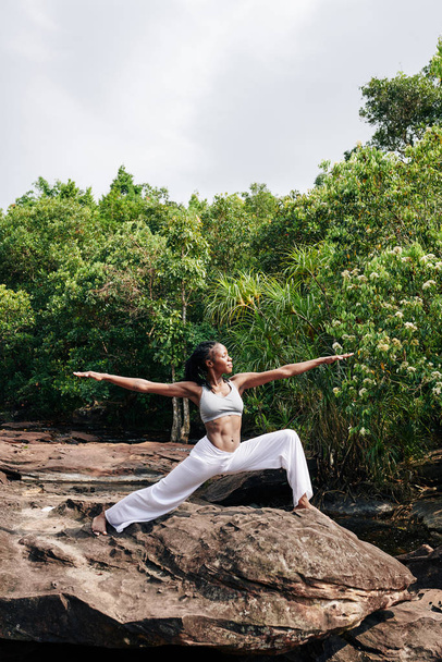 Piękna młoda czarna kobieta stojąca na dużej skale w odległym lesie w pozycji wojownika i ciesząca się sesją jogi na świeżym powietrzu - Zdjęcie, obraz
