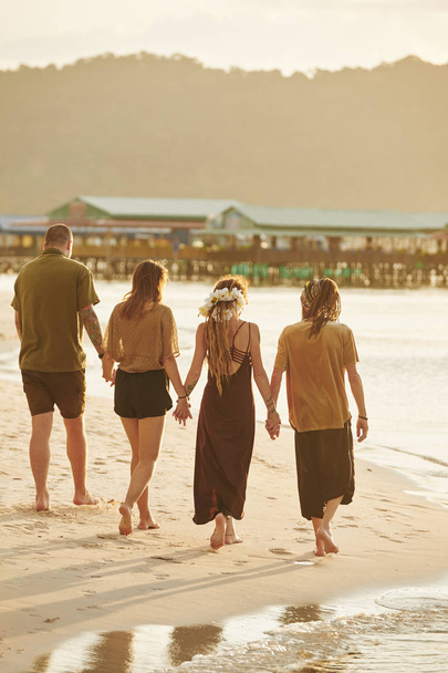 Skupina přátel držící se za ruce při procházce po pláži při západu slunce - Fotografie, Obrázek
