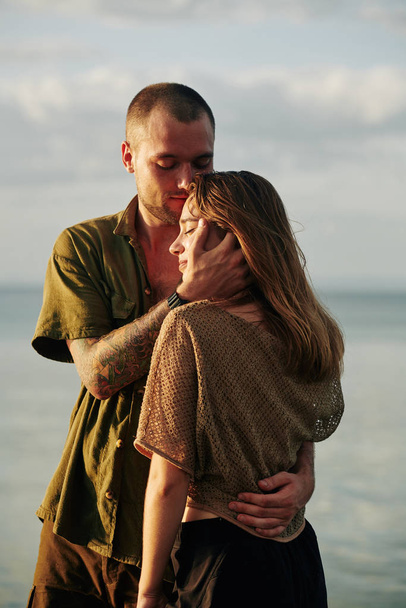 Joven abrazando y besando a su esposa en la frente con amor y ternura cuando están de pie en los rayos del sol puesta del sol
 - Foto, Imagen