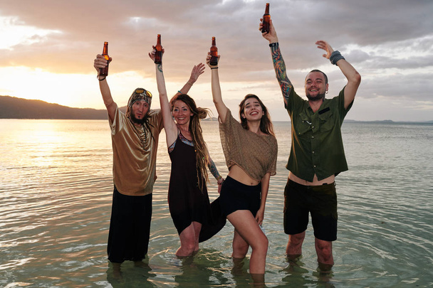 Szczęśliwi przyjaciele tańczący w wodzie i podnoszące ręce z butelkami piwa podczas zabawy na plaży - Zdjęcie, obraz