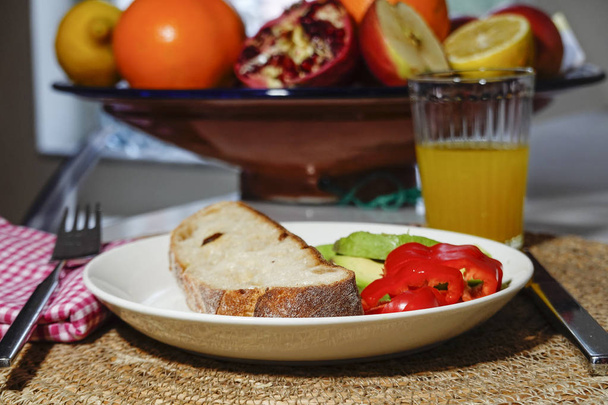 A piece of bread with avocado and peppers on a plate, with a ginger shot, and a fruit basket. - Fotoğraf, Görsel