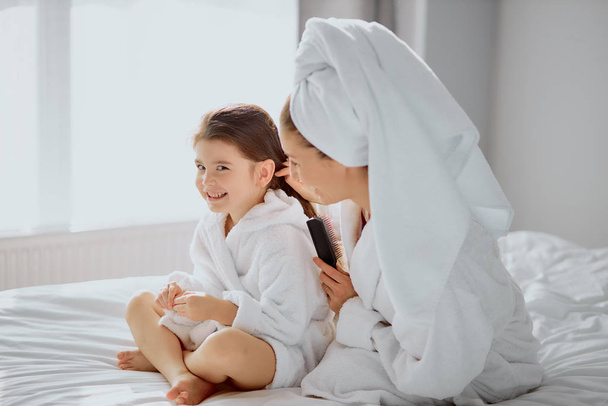 cheerful friendly family of mother and daughter combing hair on bed - Foto, Bild