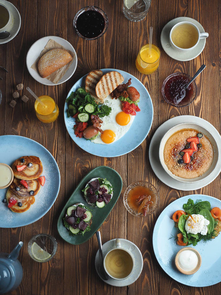 Сельский стол на завтрак в ресторане: яйца с сосисками, блины, бутерброды, кондитерские изделия и чай
 - Фото, изображение
