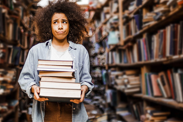 Heavy book pile in his hands, a young girl, an African-American student, looks puzzled and unhappy with tasks at the university. Stands in the library against the background of bookshelves  - Foto, Bild