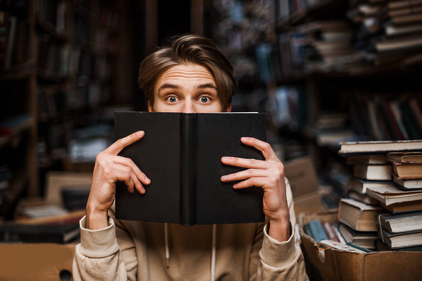 Stunned guy hiding behind a book, peeps out with surprised shocked eyes. New interesting information, Education lover surrounded by books in the library. student lifestyle. Reading time.  - Zdjęcie, obraz