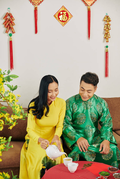 Молодая вьетнамская пара в традиционных платьях празднует Лунный Новый год и пьет травяной чай с сушеными ягодами
 - Фото, изображение