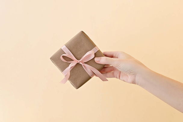 Vrouwelijke hand houden geschenkdoos verpakt in ambachtelijk papier met roze lint door op bleke beige achtergrond. - Foto, afbeelding