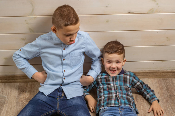 ein 4-jähriger Junge im blauen Klepy-Hemd weint auf hellem Holzgrund und sein 10-jähriger Bruder liegt auf dem Boden und hat Spaß. - Foto, Bild