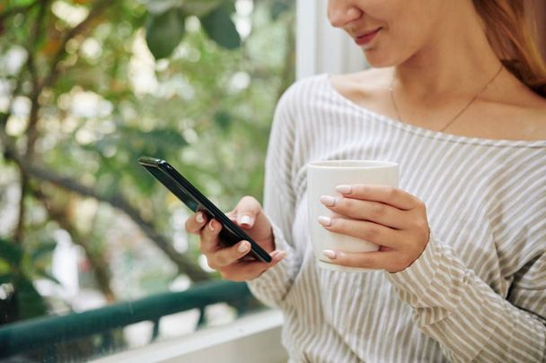 abgeschnittenes Bild einer lächelnden jungen Frau, die eine Tasse Kaffee trinkt und SMS im Smartphone checkt - Foto, Bild