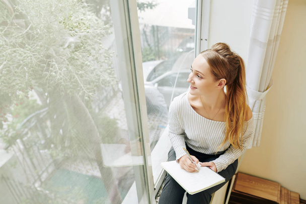 lächelnde kreative junge Frau sitzt auf Fensterbank und zeichnet Baum hinter ihrem Fenster - Foto, Bild