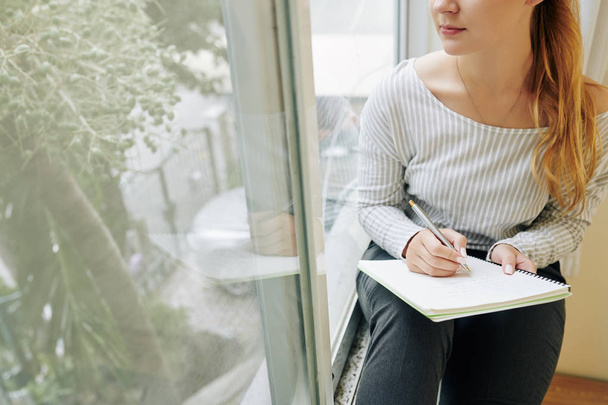 Cropped image of pensive young woman sitting on window sill and writing in diary or gratitude journal - Foto, imagen