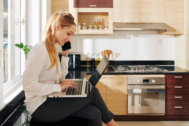 Young positive businesswoman sitting on kitchen counter, reading e-mails on laptop and talking on phone with client - Photo, Image