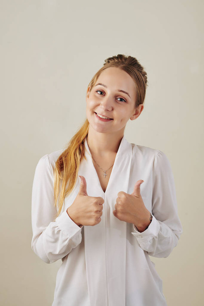 Портрет улыбающейся молодой бизнесвумен в белой блузке с большим пальцем вверх
 - Фото, изображение