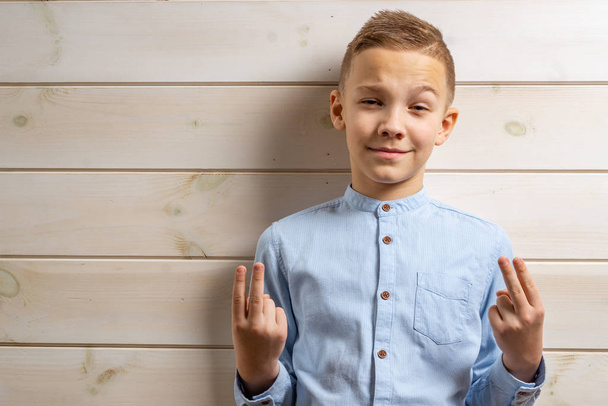 ein zehnjähriger Junge im blauen Hemd lächelt auf hellem Holzgrund und macht mit seinen Händen verschiedene Zeichen. - Foto, Bild