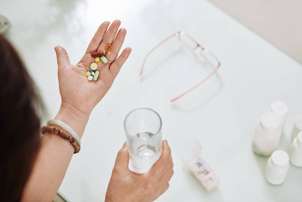 Руки женщины с суточной дозой различных таблеток и добавок и стаканом пресной воды
 - Фото, изображение
