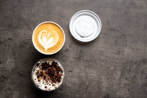 Toetje in plastic bakje en kopje cappuccino bovenaanzicht op grijze achtergrond - Foto, afbeelding