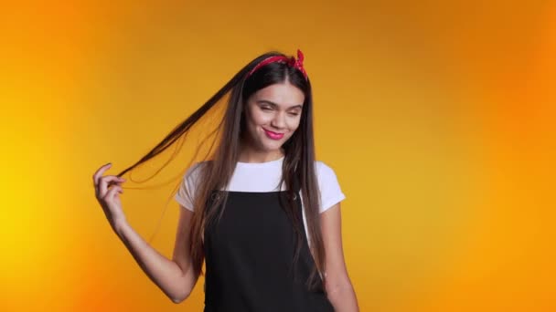 Pretty young girl with long hair standing on yellow studio background, smiling and flirting. Cute portrait of woman in black overall - Záběry, video