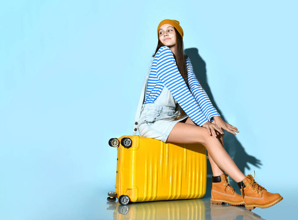 Teenager in jeans overall skirt, striped sweatshirt, boots and hat. She sitting on yellow suitcase, blue background. Full length - Фото, зображення