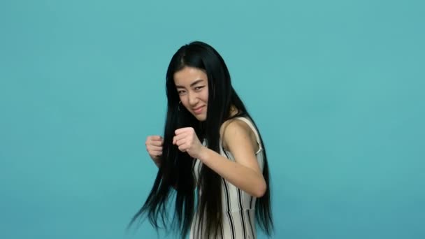 Let's fight! Aggressive asian woman with long straight black hair in dress ready to boxing with clenched fists, threatening to punch, fighting spirit. indoor studio shot isolated on blue background - Footage, Video