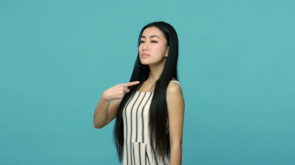 Stunning arrogant asian woman with long straight black hair pointing herself and looking with haughty expression, proud of her beauty and success, self-love concept. indoor studio shot blue background - Felvétel, videó