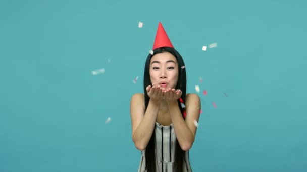Happy birthday! Joyful asian woman with long black hair with funny party cone on head blowing confetti from hands and smiling, party celebration. indoor studio shot isolated on blue background - Video