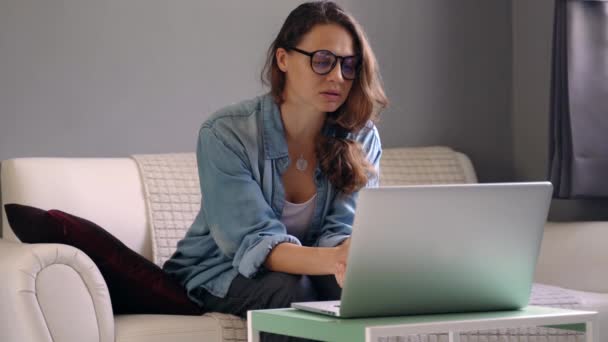 A young woman with a laptop sitting on a sofa and holds an online consultation. - Imágenes, Vídeo