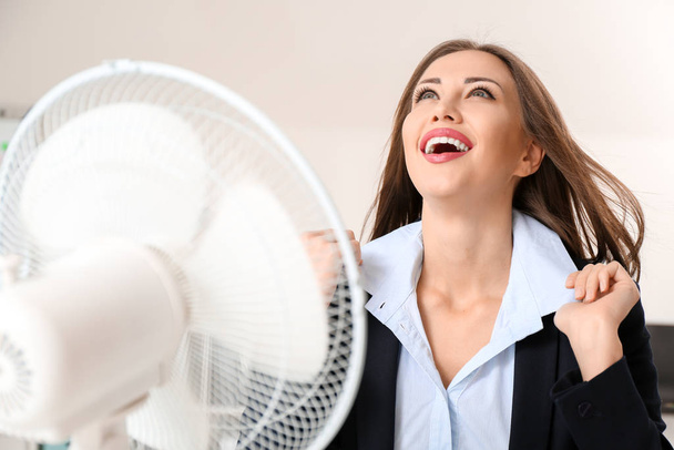 Young woman using electric fan during heatwave in office - Fotoğraf, Görsel
