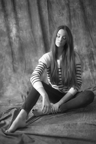 Lovely young girl model with dark long hair in a striped sweater and skinny jeans posing in the studio on a textile background. Beauty and fashion. - Φωτογραφία, εικόνα