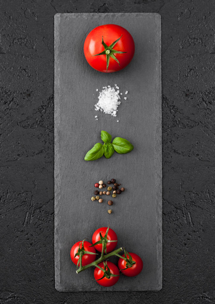 Black stone board with tomatoes, salt and pepper and basil on black table background. - 写真・画像