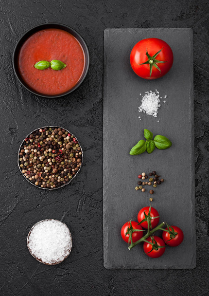 Black bowl plate of creamy tomato soup on black table background with stone chopping board and raw tomatoes, pepper and salt. - Foto, Imagem