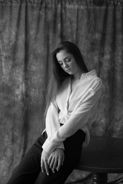 Lovely young girl model with dark long hair in a white shirt and black pants posing in the studio on a textile background. Beauty and fashion. - Foto, imagen