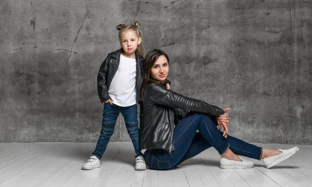 Positive young woman and little girl in similar clothing jeans and black leather jackets posing over grey concrete background - Foto, imagen