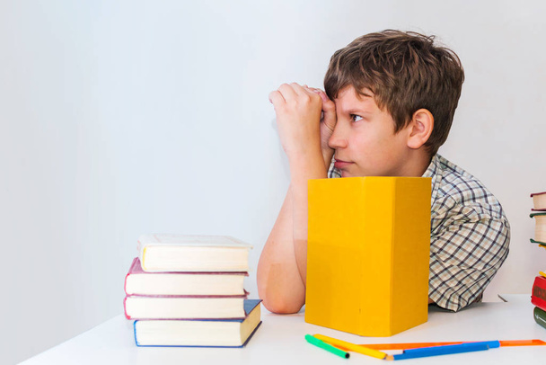 Ένα έφηβο αγόρι γκριμάτσες σε ένα τραπέζι με βιβλία που κάνει την εργασία - Φωτογραφία, εικόνα