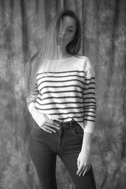Lovely young girl model with dark long hair in a striped sweater and skinny jeans posing in the studio on a textile background. Beauty and fashion. - Foto, Imagem