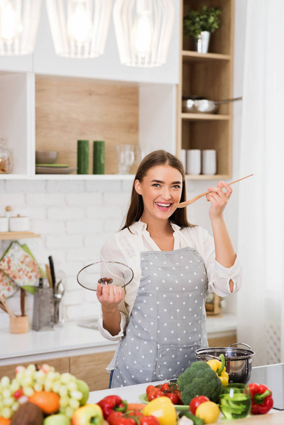 Happy woman in apron holding wooden spoon and smiling - Photo, Image