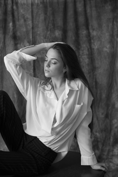 Lovely young girl model with dark long hair in a white shirt and black pants posing in the studio on a textile background. Beauty and fashion. - 写真・画像