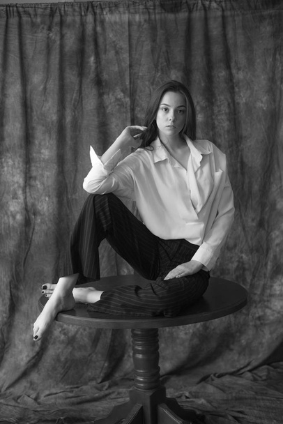 Lovely young girl model with dark long hair in a white shirt and black pants posing in the studio on a textile background. Beauty and fashion. - Zdjęcie, obraz